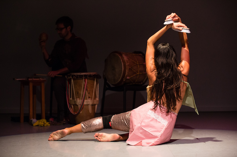 Angelica sits facing her drummer. Bother her legs are tucked under her left side. Her hands raise over her head with two scrub brushes in her hands